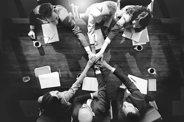 Decorative photo of six people around a table seen from above joining hands in the middle.
